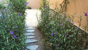 Ein gepflasterter Weg, gesäumt von üppigem Grün und violetten Blüten, führt zu einem Hauseingang.