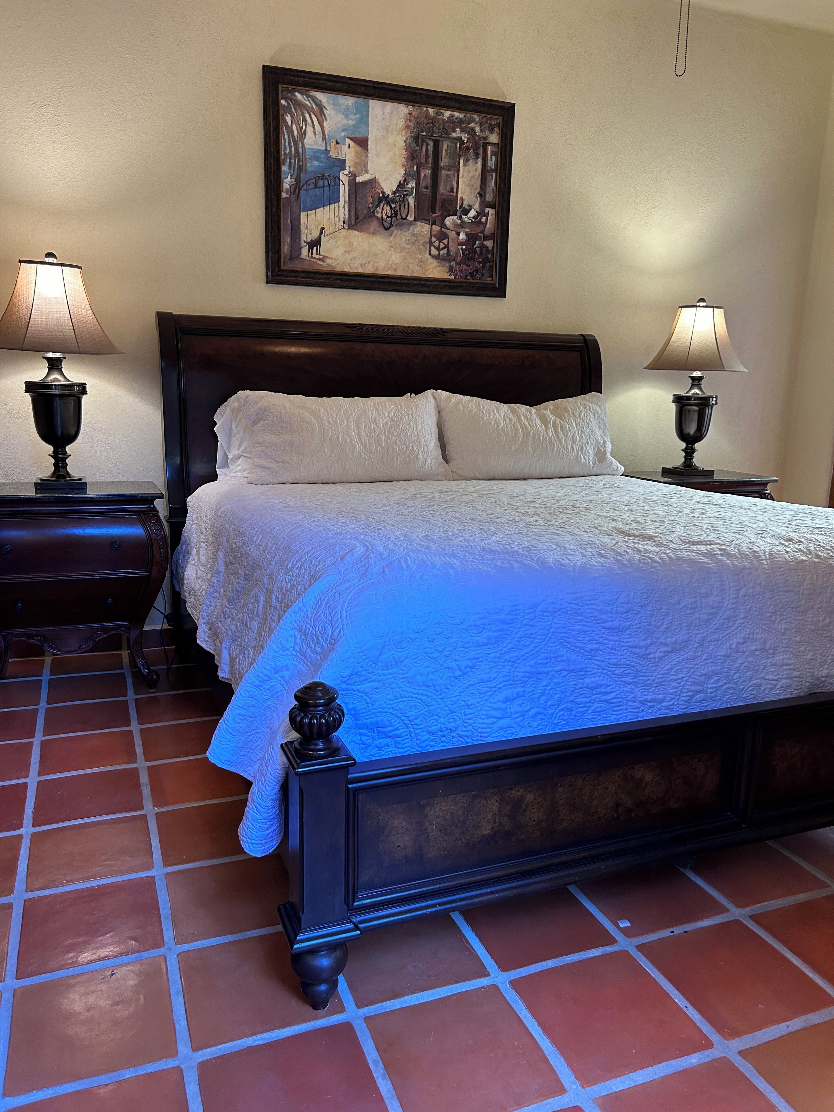 Una cama bien hecha con edredón blanco, dos mesitas de noche con lámparas, un cuadro en la pared y suelo de baldosas.