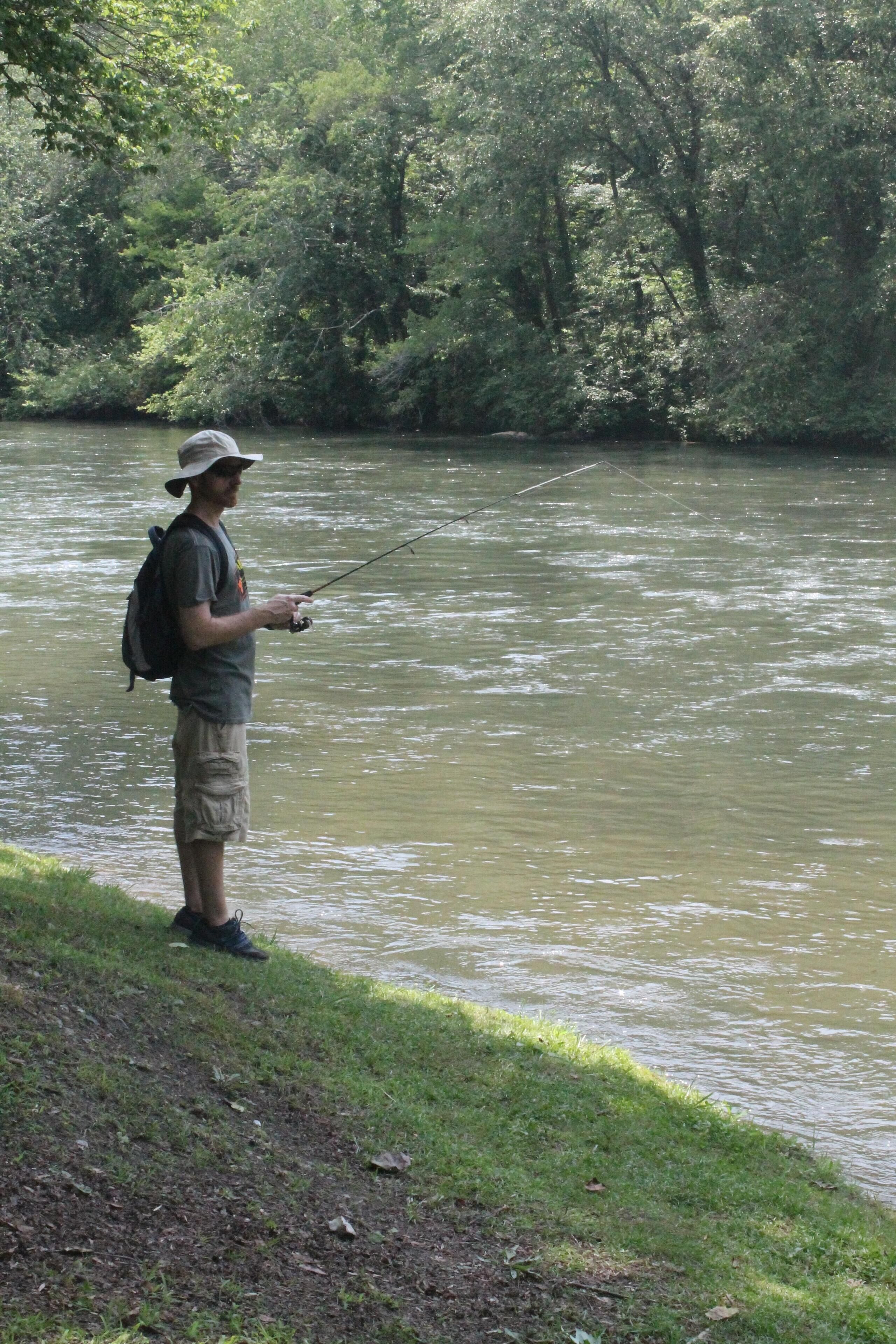 A person fishing by a river with dense greenery in the background.