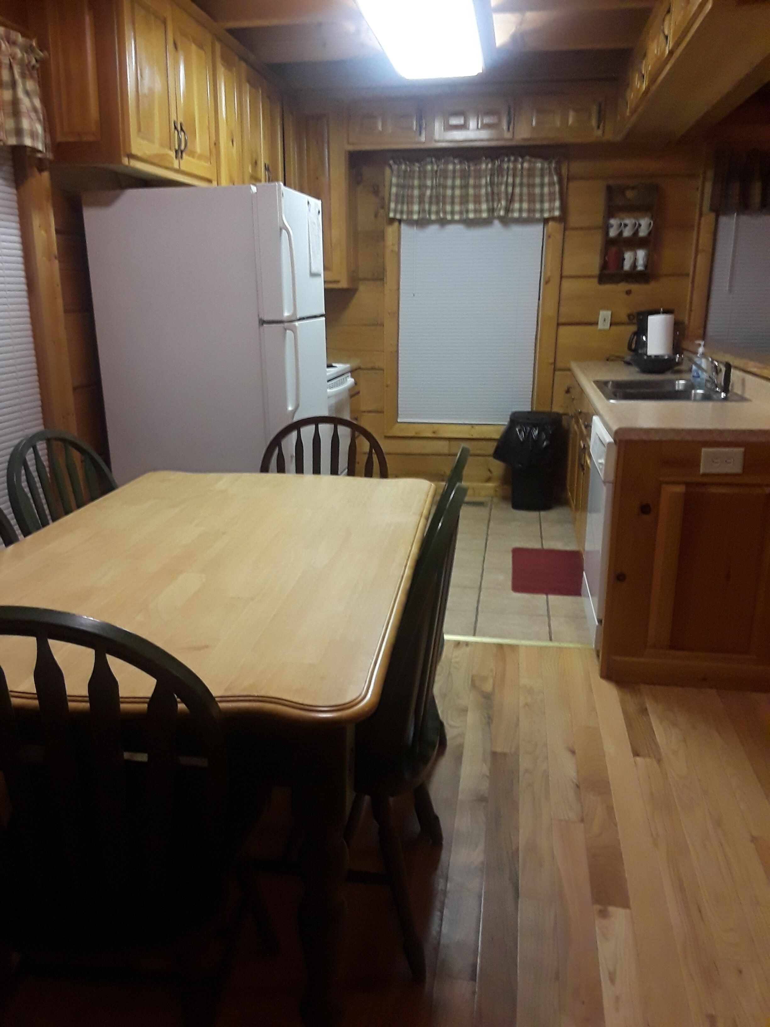 Una cocina estilo cabaña de madera con una mesa de comedor y sillas, un refrigerador, una estufa y una ventana con cortina a cuadros.