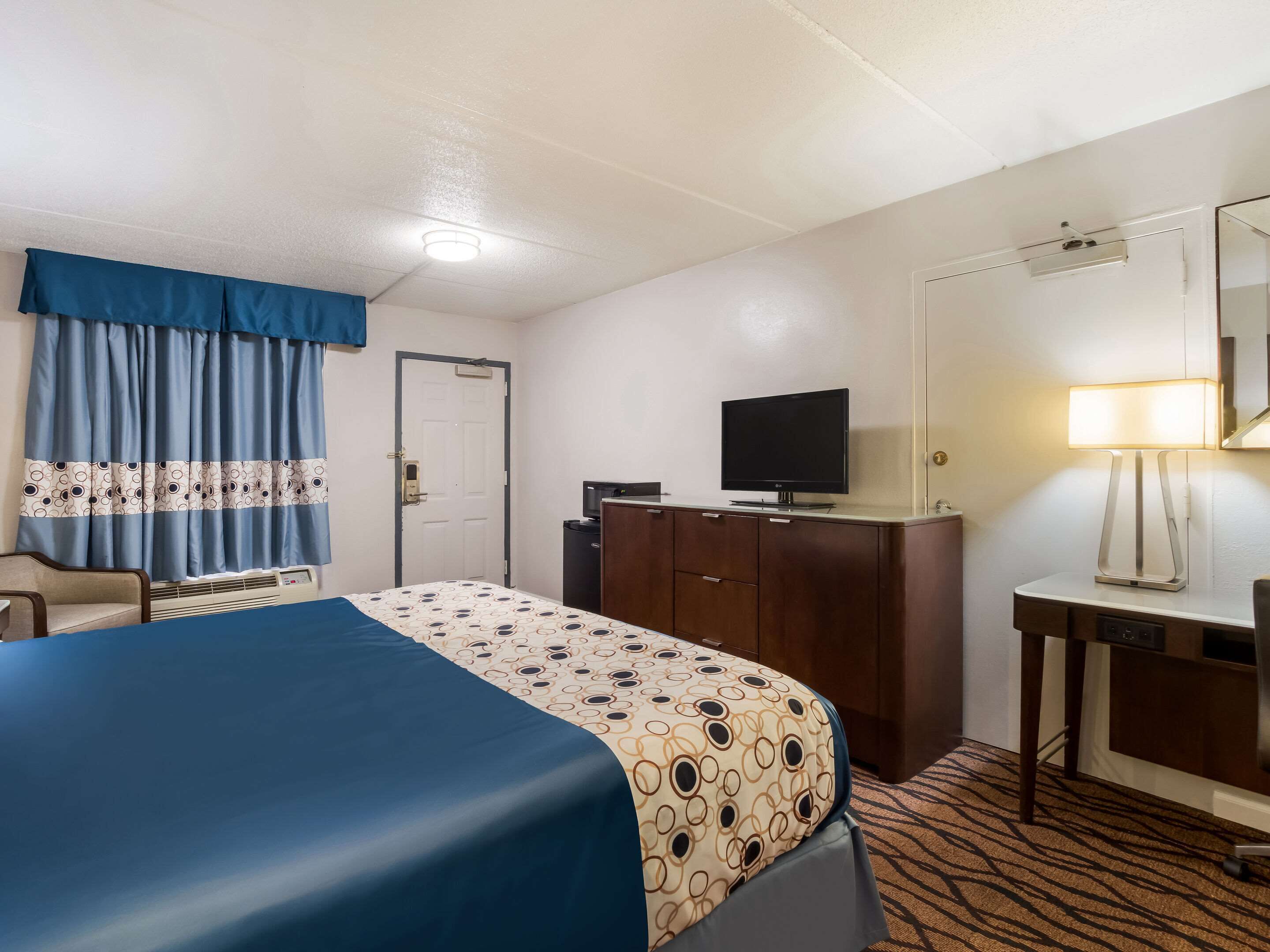 A hotel room with a bed, a desk, a television, a lamp, and a door.