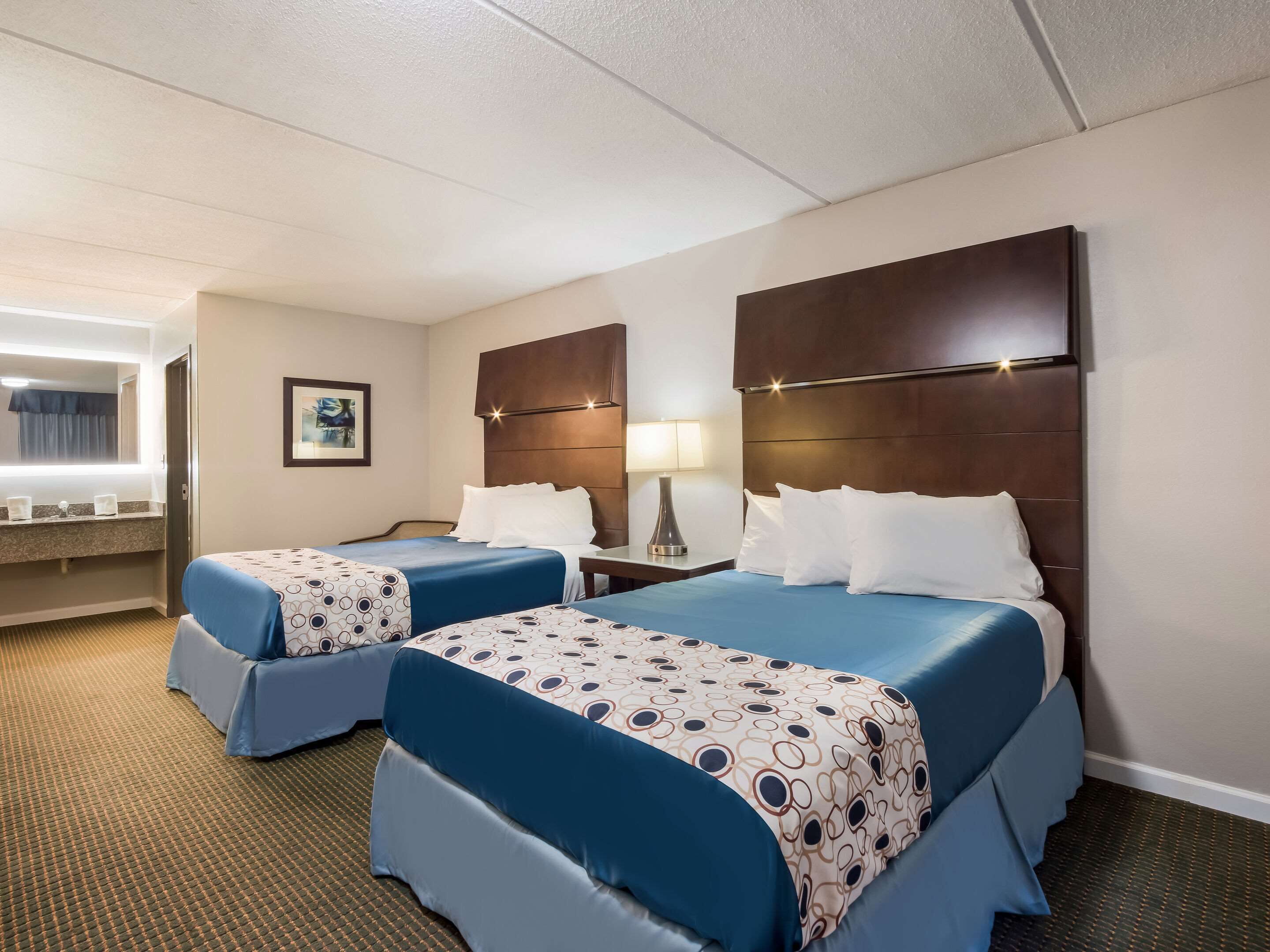 A hotel room with two beds, a nightstand with a lamp, a bathroom with a mirror and sink, and a framed picture on the wall.