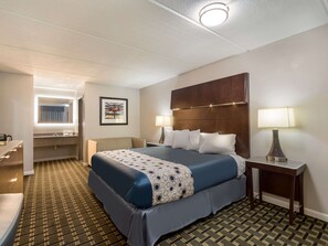 Quarto de hotel com uma cama grande, mesa de cabeceira com abajur, banheiro com espelho e pia, e carpete estampado.