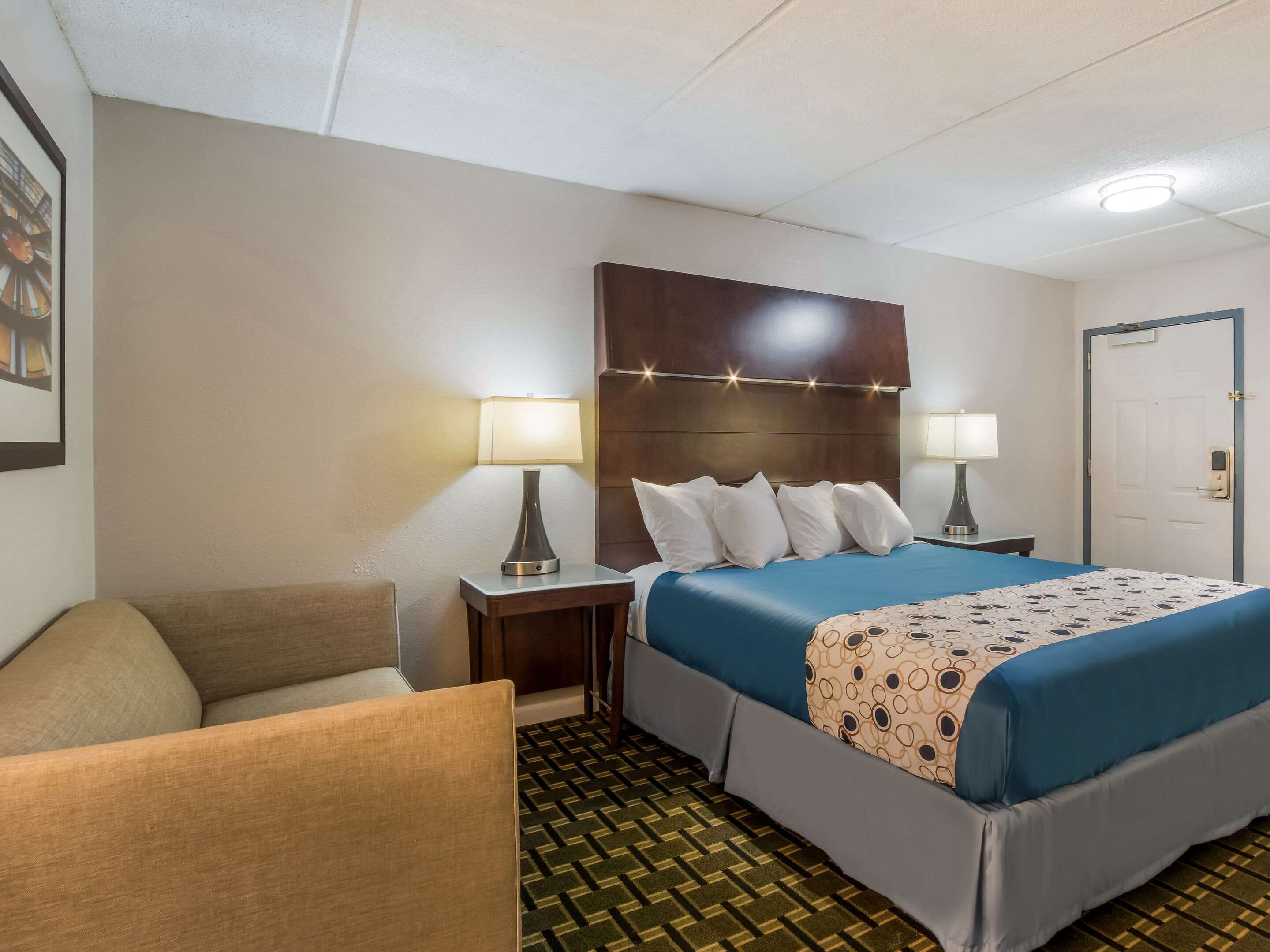 Une chambre d’hôtel avec un lit, deux tables de chevet avec des lampes, une chaise, un tapis à motifs et une porte.