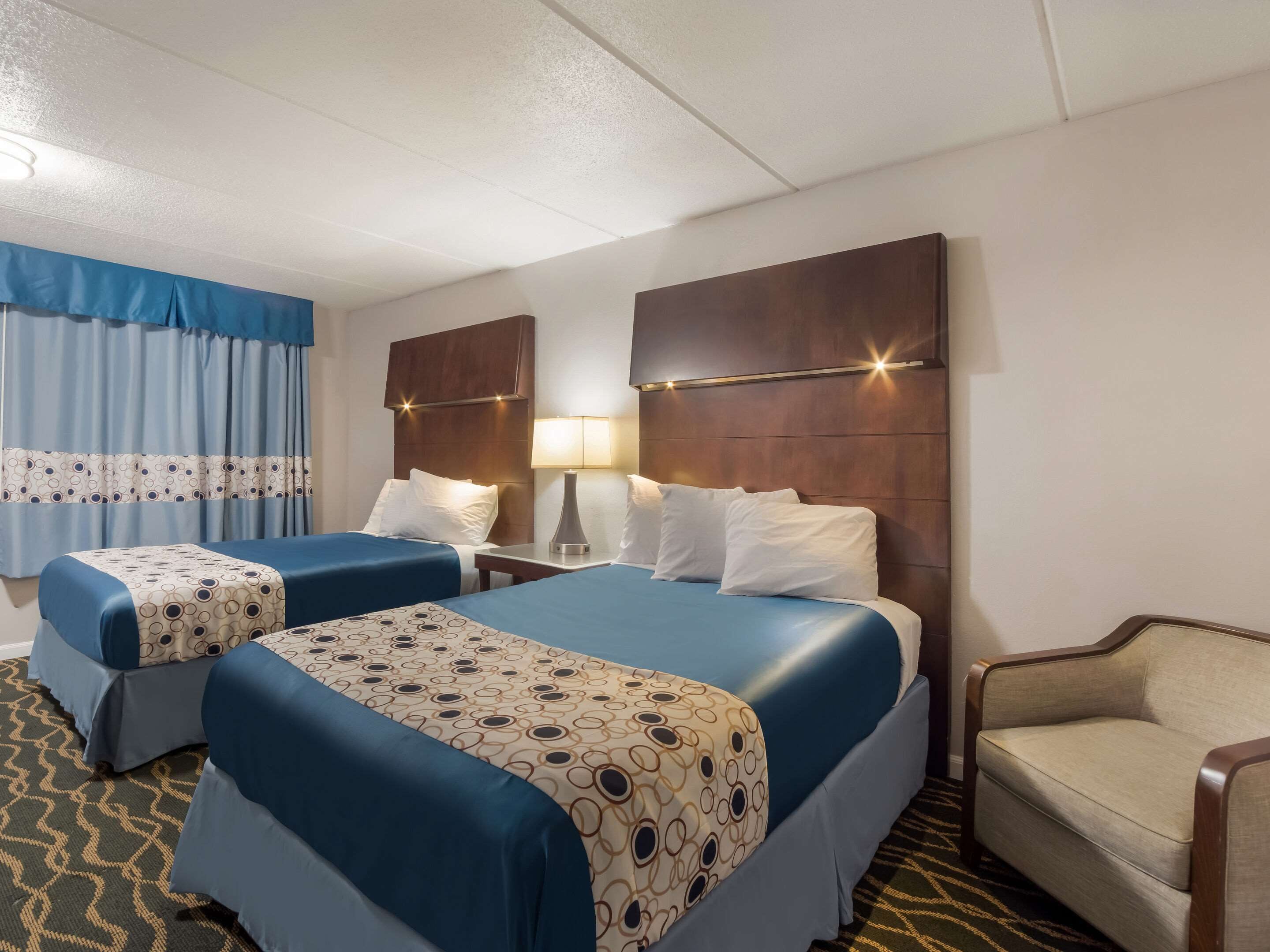 Ein Hotelzimmer mit zwei Betten, einem Schreibtisch, einem Sessel und einem gemusterten Teppich.