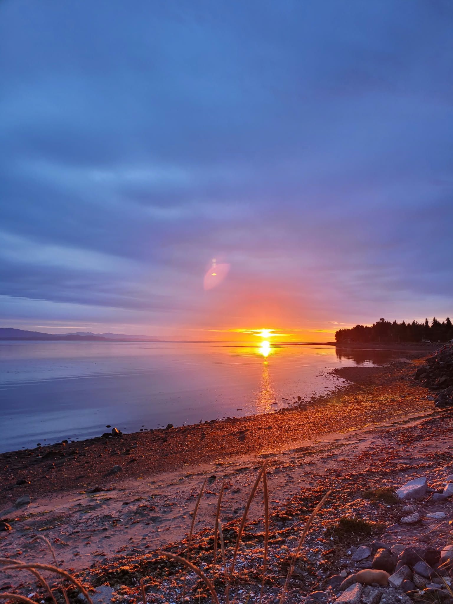 Un atardecer sobre una superficie de agua tranquila con una costa rocosa y una zona boscosa en la distancia.
