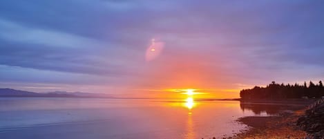 Un atardecer sobre una superficie de agua tranquila con una costa rocosa y una zona boscosa en la distancia.