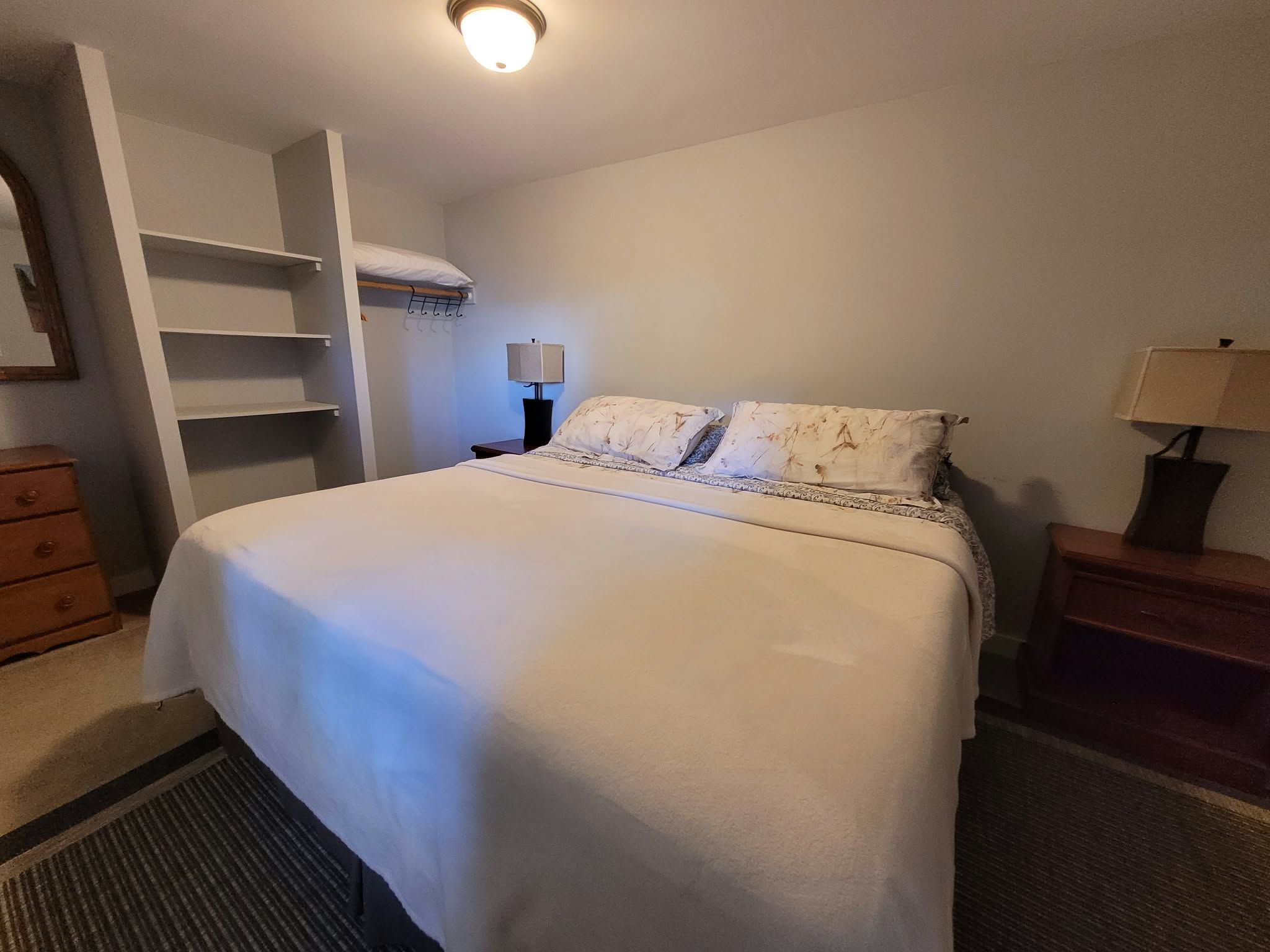 Hotellihuone, jossa on sänky, yöpöytä, lamppu, vaatekaappi ja lipasto.