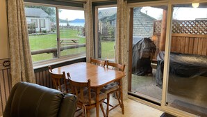 A dining area with a wooden table and chairs, large windows with curtains, and a view of a backyard with a wooden deck and picnic table. - Seaside Beach Croft Cabin (Qualicum Beach)