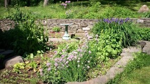 A garden with a stone wall, a birdbath, and various plants including purple flowers. - Historic 6-bedroom manor next to Beaverkill River — sleeps 18, 5 en-suite baths. (East Branch)