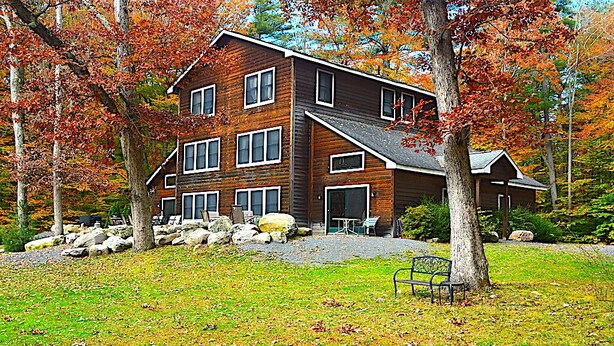 A multi-story wooden cabin with a stone foundation, surrounded by a grassy area and autumn-colored trees.