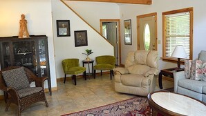 A living room with a staircase, a cabinet with glass doors, two green armchairs, a beige armchair, a patterned rug, and a table.