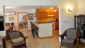 A spacious living room with two wicker chairs, a dining area with a table and chairs, and a kitchen with wooden cabinets and a central island.