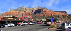 Une rue bordée de voitures garées et où circule un autobus, des immeubles avec des enseignes, et une montagne en arrière-plan.