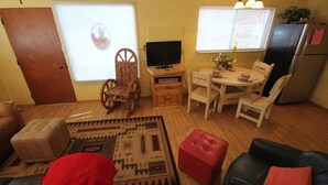 Een knus woongedeelte met een bank, een rode poef, een eettafel met stoelen, een televisie op een houten dressoir en een koelkast.