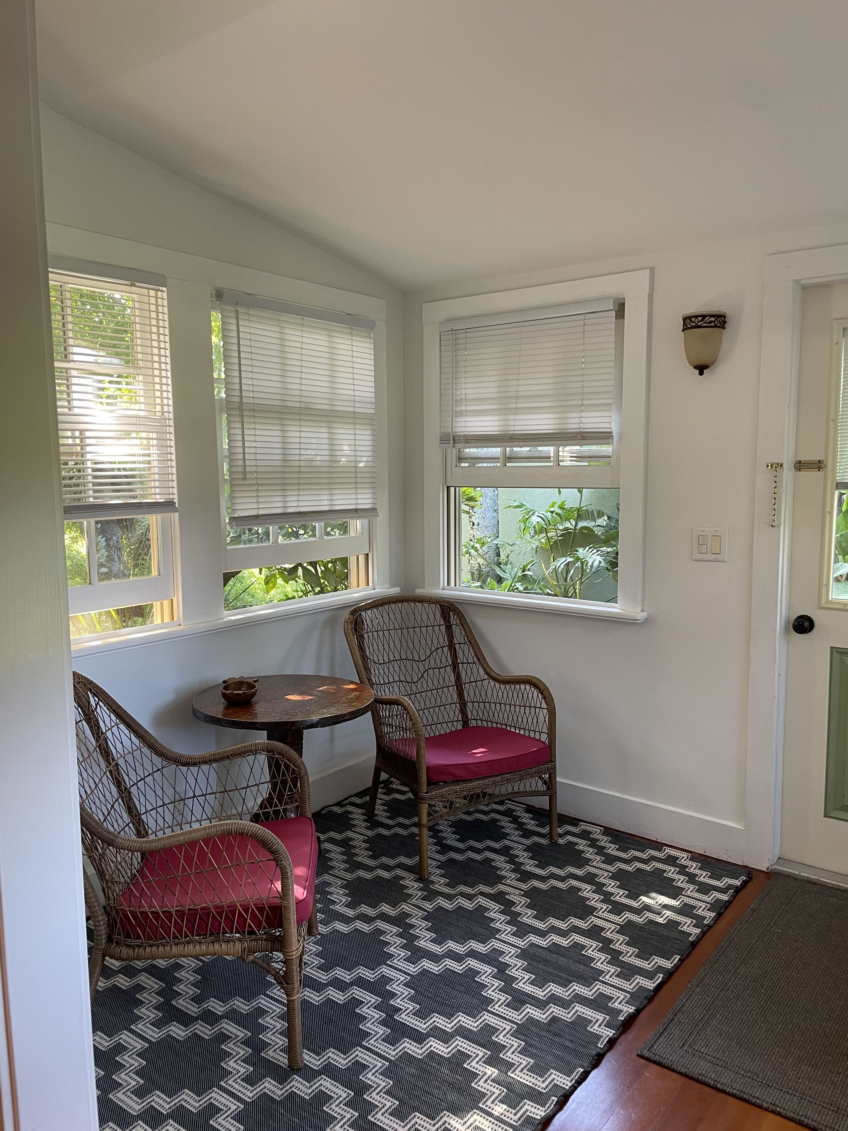A corner room with a bay window, wicker chairs, a small table, and a patterned rug.
