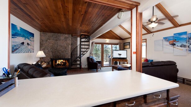 A spacious living area with a stone fireplace, wooden ceiling beams, a large white table, and a staircase leading to a balcony.