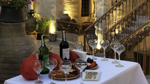 Ein Tisch ist gedeckt mit Weingläsern, Flaschen und Speisen in einem von Steinmauern umgebenen Innenhof mit Bögen und einer Treppe.