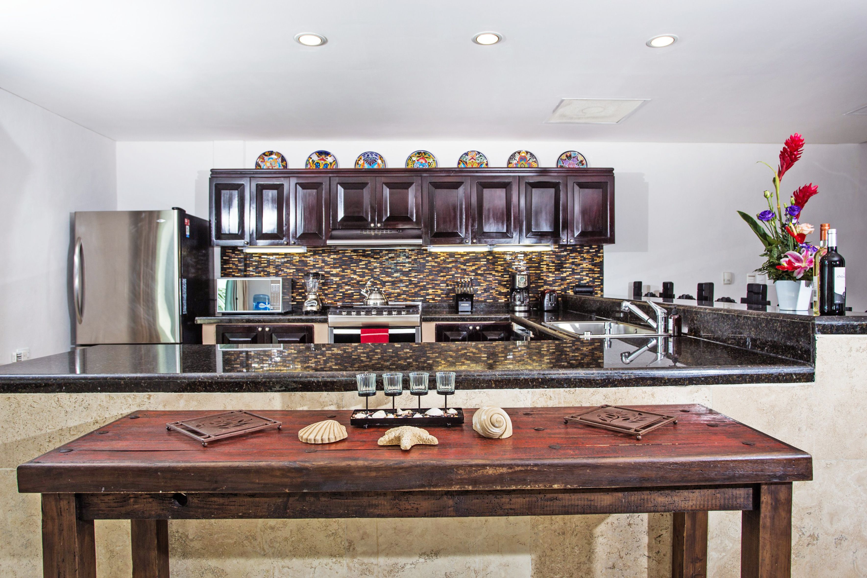 A modern kitchen with a stainless steel refrigerator, dark wooden cabinets, a tiled backsplash, and a wooden table with decorative shells.