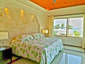 Ein Schlafzimmer mit einem Stein-Kopfteil, einem Bett mit einem blattgemusterten Bettüberwurf, einem Nachttisch mit einer Lampe und Blick auf Palmen und das Meer durch ein großes Fenster.
