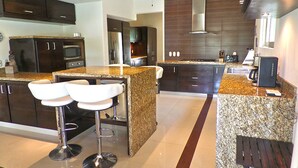 Eine moderne Küche mit dunklen Holzschränken, Granit Arbeitsplatten und Edelstahlgeräten. Zwei weiße Barhocker und eine kleine Holzbank sind ebenfalls vorhanden.