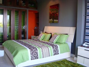 Een moderne slaapkamer met een groot bed, beddengoed in groen en bruin, een nachtkastje en een schilderij van een boom.