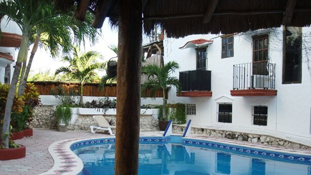Una piscina con borde de azulejos, rodeada de palmeras y un edificio blanco con balcones.