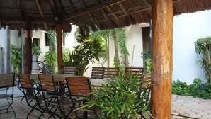Un patio al aire libre con mesas y sillas de madera, rodeado de plantas en macetas y un techo de paja.
