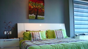 Un dormitorio con una cama, ropa de cama verde, una mesita de noche, una lámpara y un cuadro de un árbol.