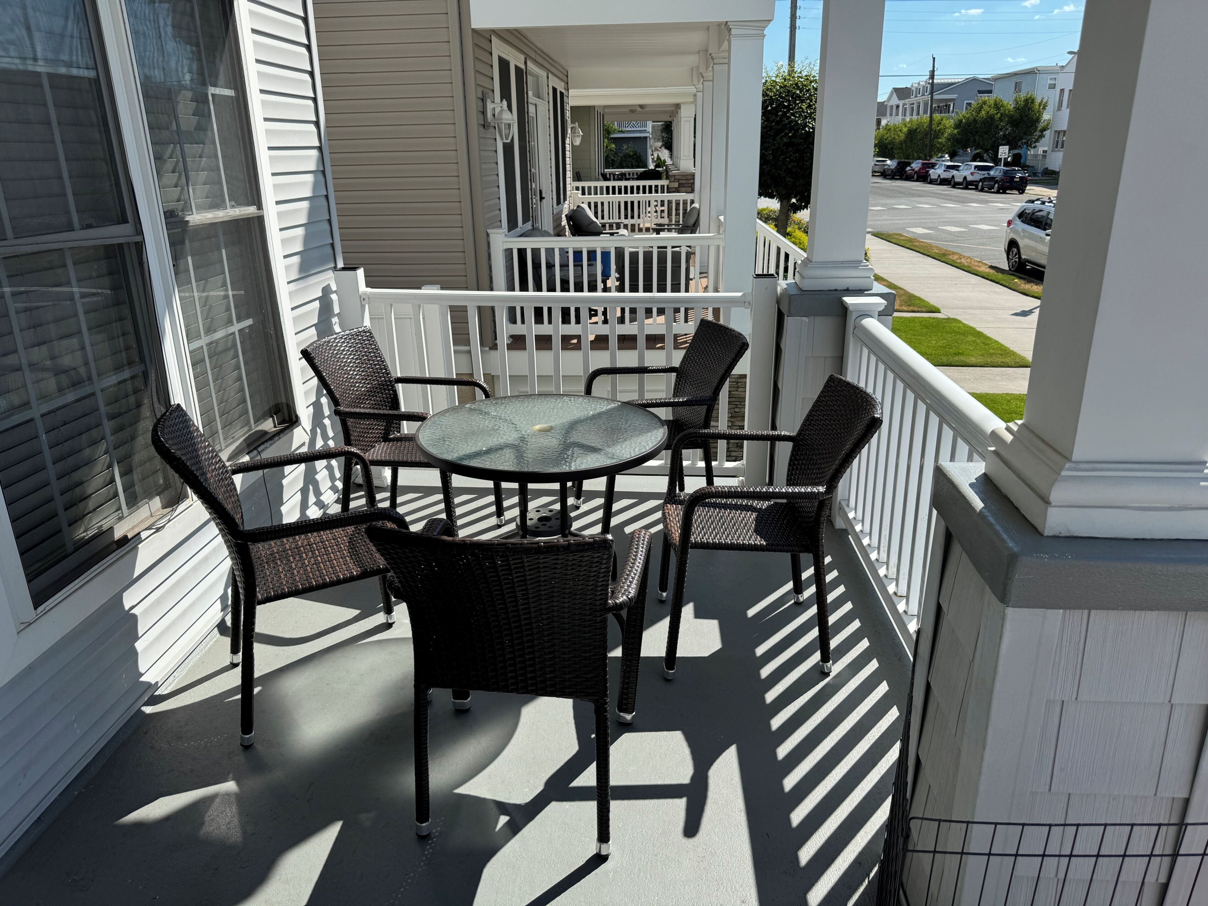 A covered patio with a round table and four chairs, a white railing, and a view of a street with parked cars.