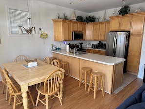 A kitchen with wooden cabinets, a stainless steel refrigerator, a microwave, and a sink. There is a dining table with chairs and a bar stool.