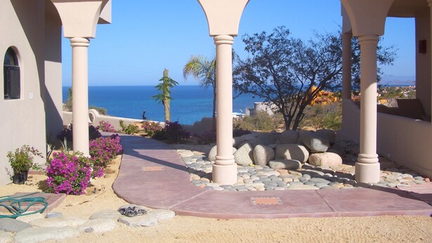 A covered outdoor area with a view of the sea, featuring a stone pathway, archways, and some landscaping.