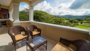 Un balcón con mobiliario de mimbre y una mesa de vidrio, con vistas a un paisaje exuberante con árboles y colinas al lejano.