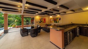 A modern kitchen with a bar area, dining space with wicker furniture, and a view of lush greenery outside. - Los Suenos Luxury Villas by Stay in Costa Rica (Los Sueños)