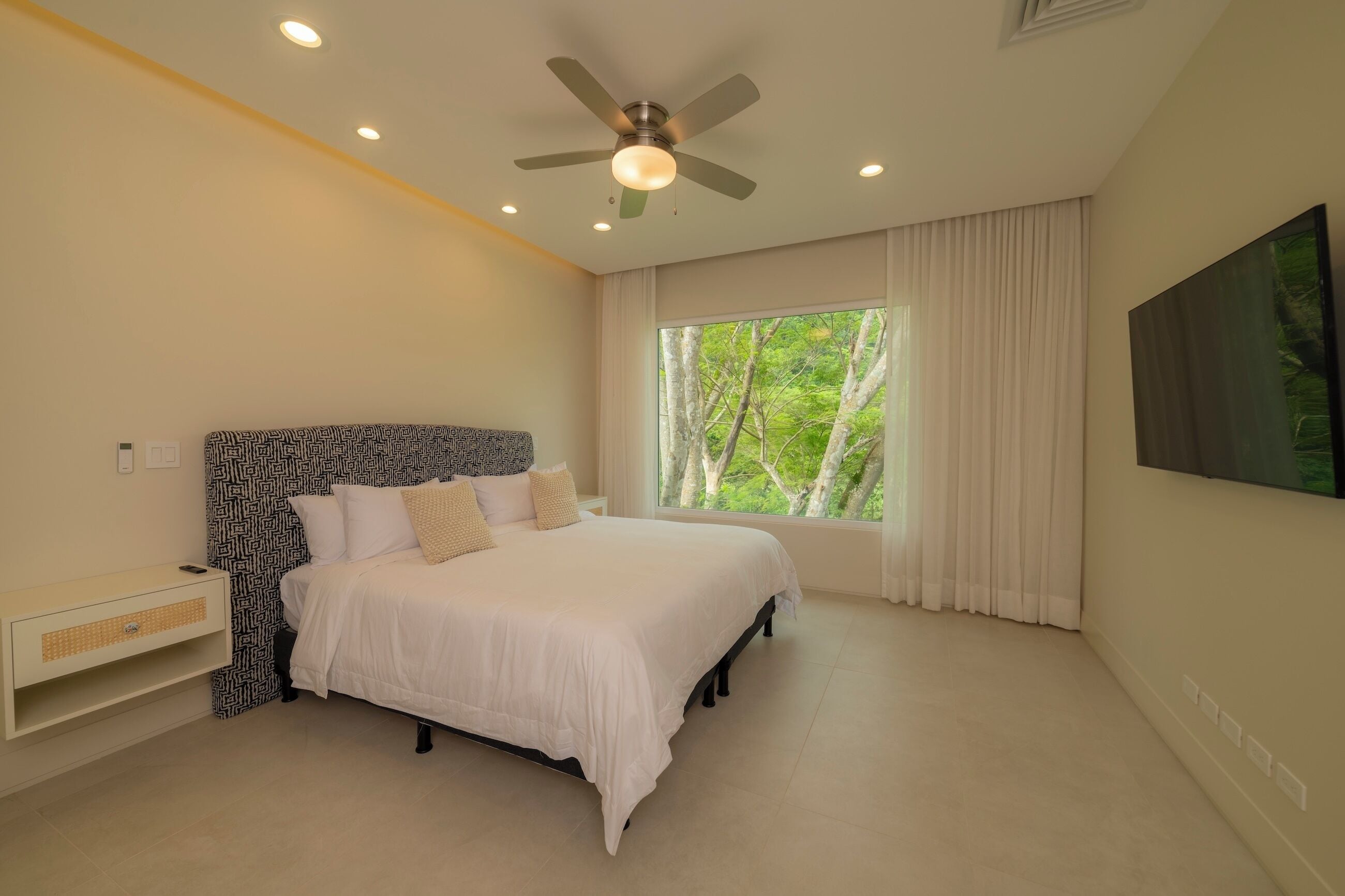 Quarto com cama, ventoinha no teto, janela com cortinas e televisão na parede.