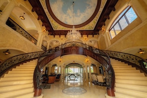 A grand staircase with ornate railings and a chandelier, leading to a spacious lobby with a high ceiling and a skylight.