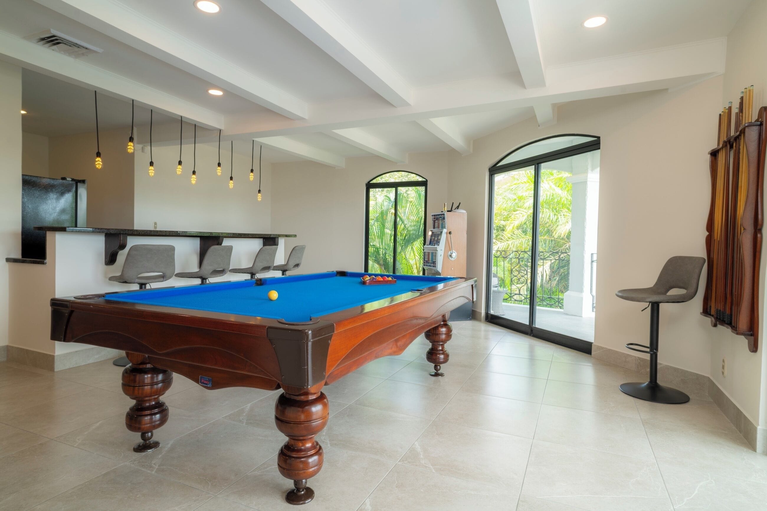 A pool table in a room with a modern design, a bar area, and a view of greenery through large windows.