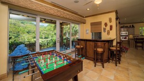 A room with a foosball table, a bar area, and large windows offering a view of greenery.
