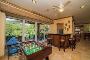 A room with a foosball table, a bar area, and large windows offering a view of greenery.