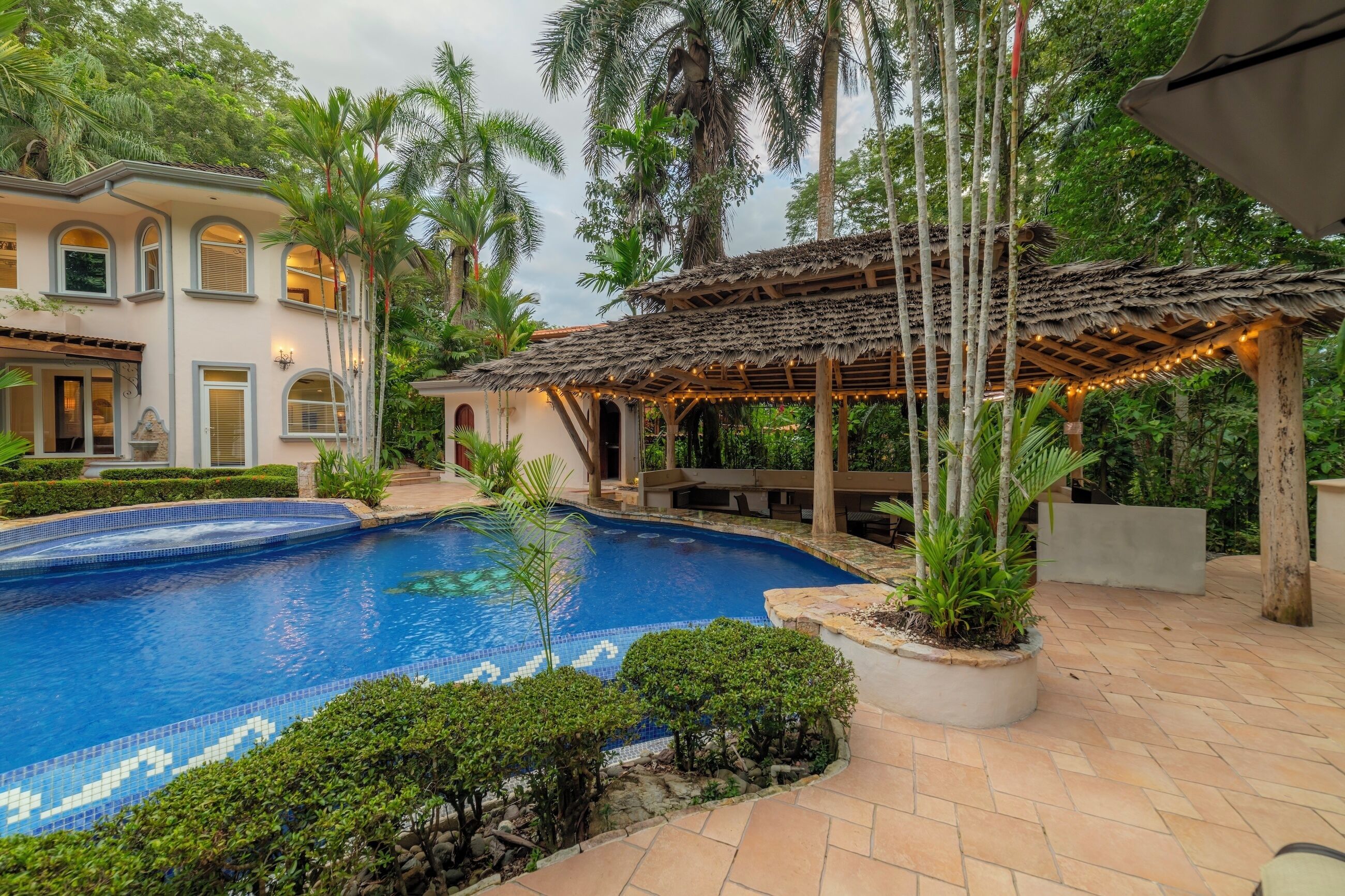 Piscina com borda em azulejo, rodeada por um jardim ajardinado com diversas plantas e uma pérgula com luzes de cordão.