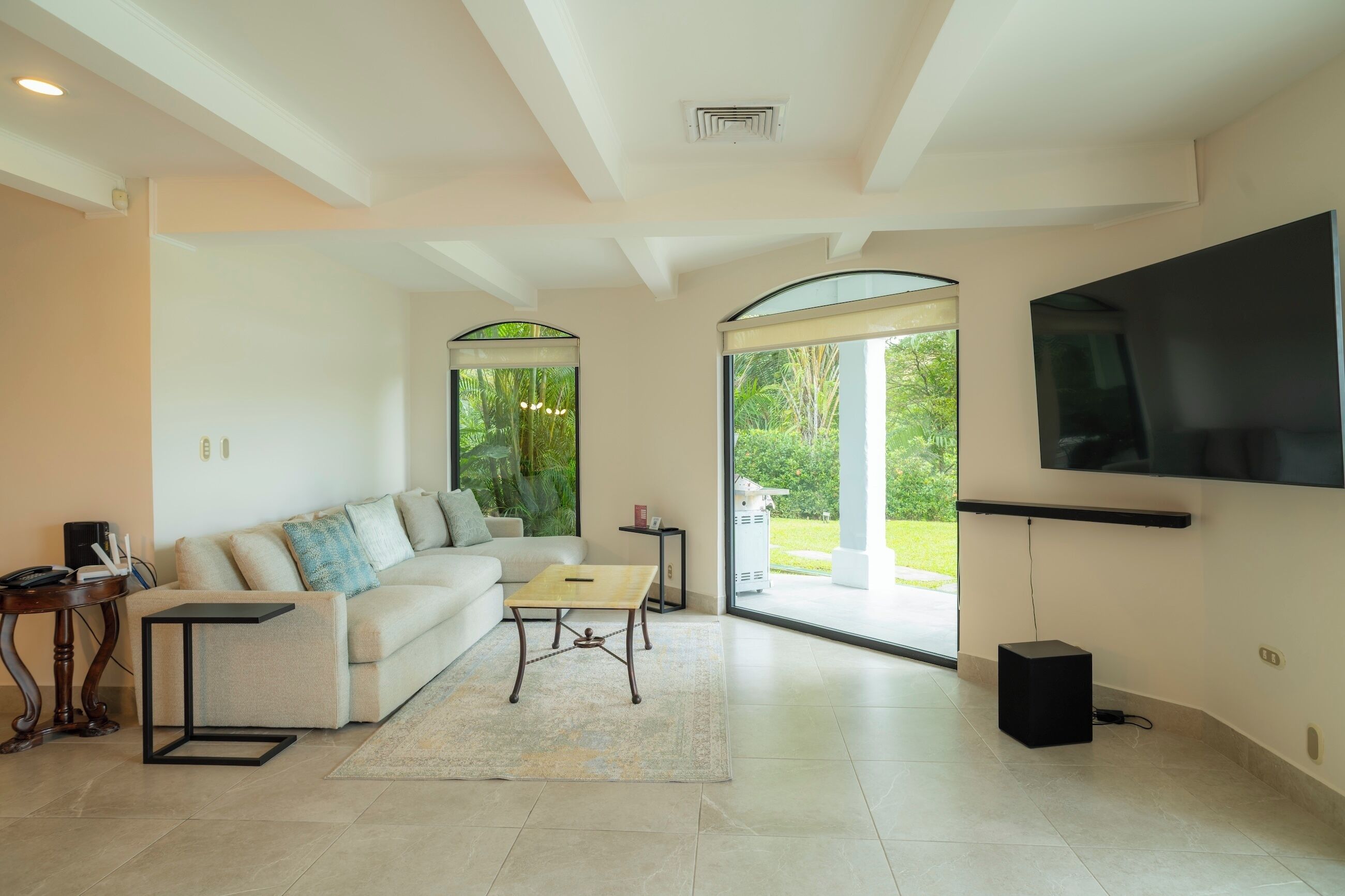 Uma sala de estar moderna com uma grande televisão de tela plana, um sofá, uma mesa de centro e vista para um jardim através de uma porta de vidro.