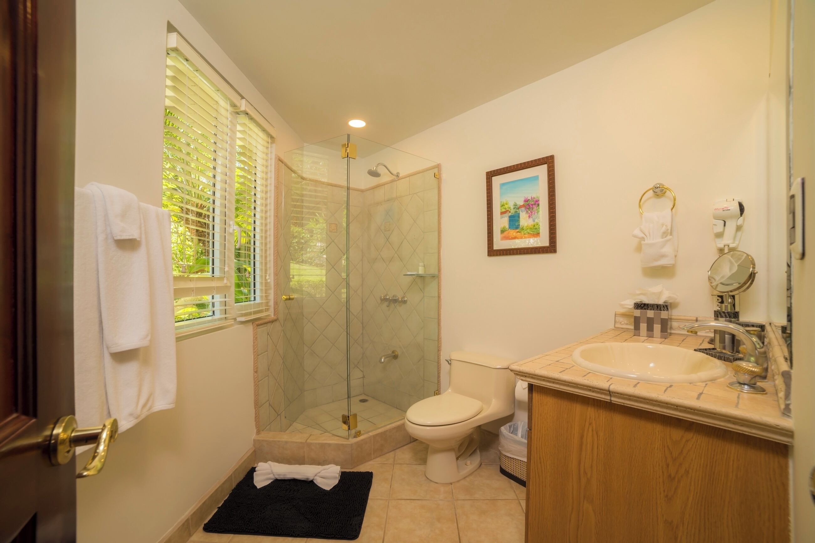 A bathroom with a glass-enclosed shower, a toilet, a sink with a mirror, and a window with blinds.