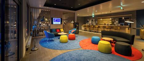 A modern hotel lobby with a bar area, seating with ottomans, and a television screen displaying hotel information.