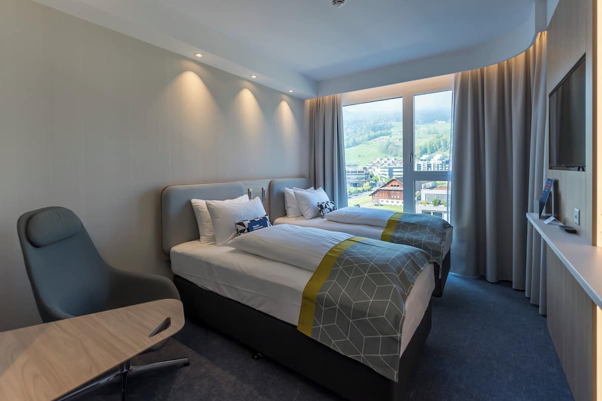 Een hotelkamer met twee bedden, een bureau, een stoel, een televisie en een groot raam met uitzicht op een stad.
