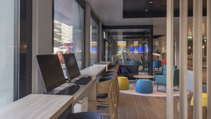 Un espace de travail moderne doté de plusieurs bureaux et chaises, de grandes fenêtres et d’un coin salon avec des chaises et des tables aux couleurs vives.