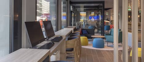 A modern workspace with multiple desks and chairs, large windows, and a seating area with colorful chairs and tables.