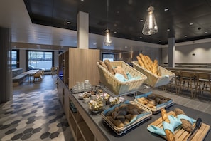 A modern breakfast buffet with wicker baskets of bread, a variety of pastries, and a seating area with tables and chairs. - Holiday Inn Express Luzern - Kriens by IHG (Kriens)