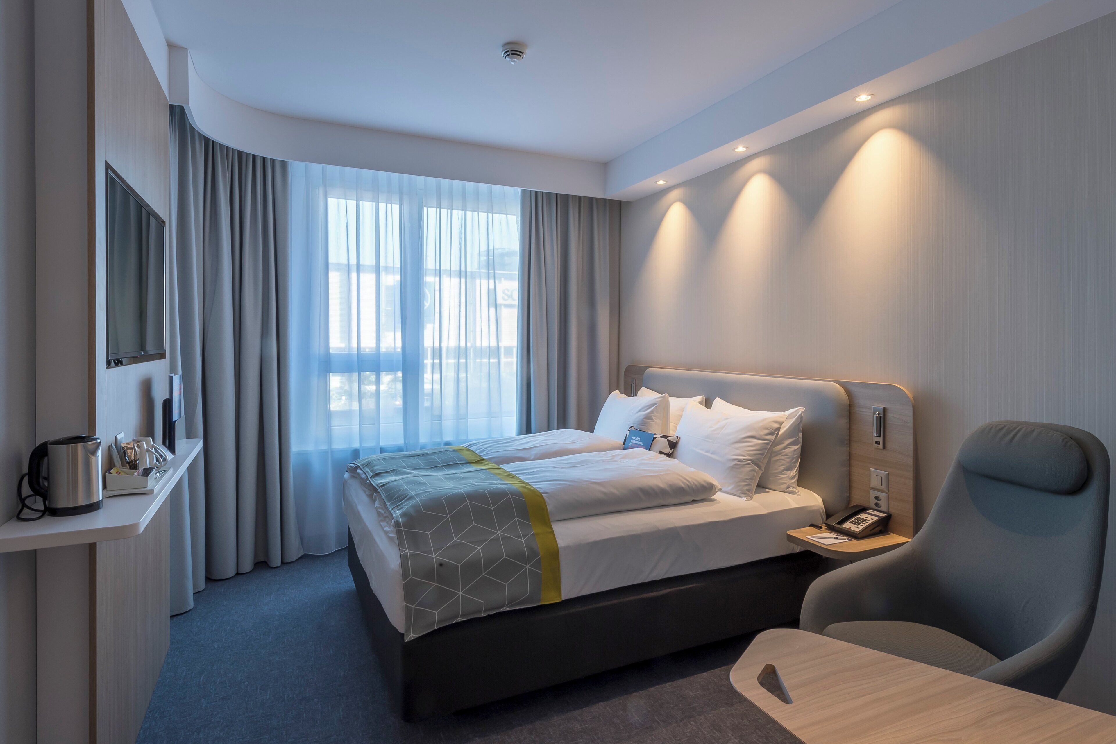 Una habitación de hotel con cama, mesita de noche, silla, escritorio y una ventana grande con cortinas.