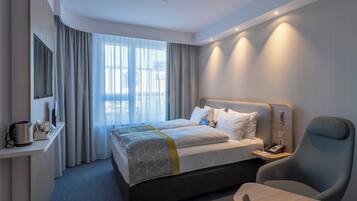 Una habitación de hotel con cama, mesita de noche, silla, escritorio y una ventana grande con cortinas.
