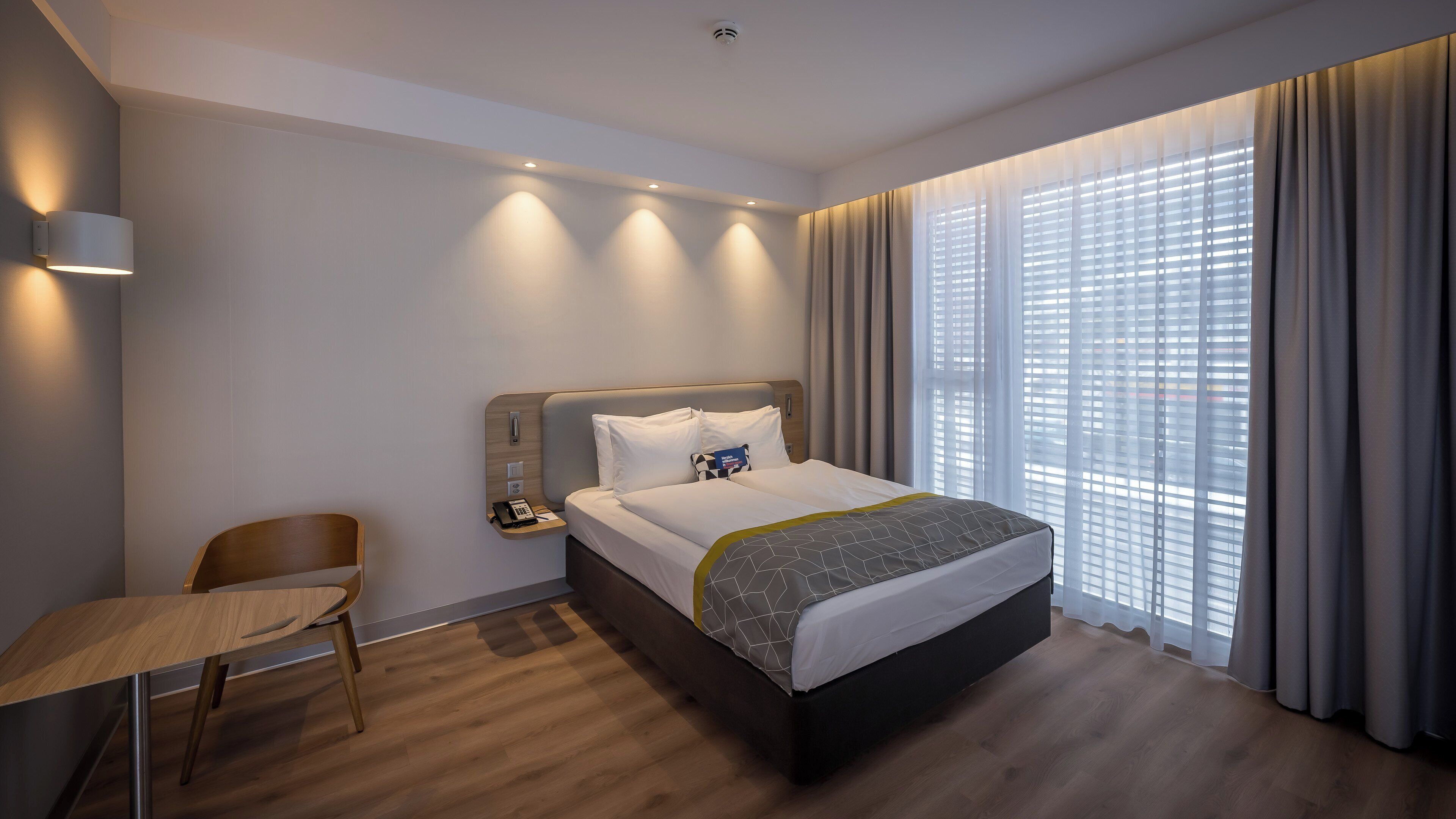 Une chambre d’hôtel moderne avec un grand lit, une table de chevet avec un téléphone et un livre, une chaise et une grande fenêtre avec des rideaux.
